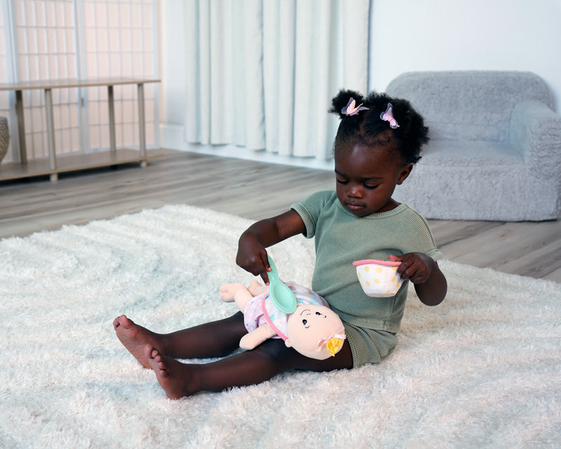 toddler age girl seated, pretending to feed soft baby doll with pretend spoon and food bowl