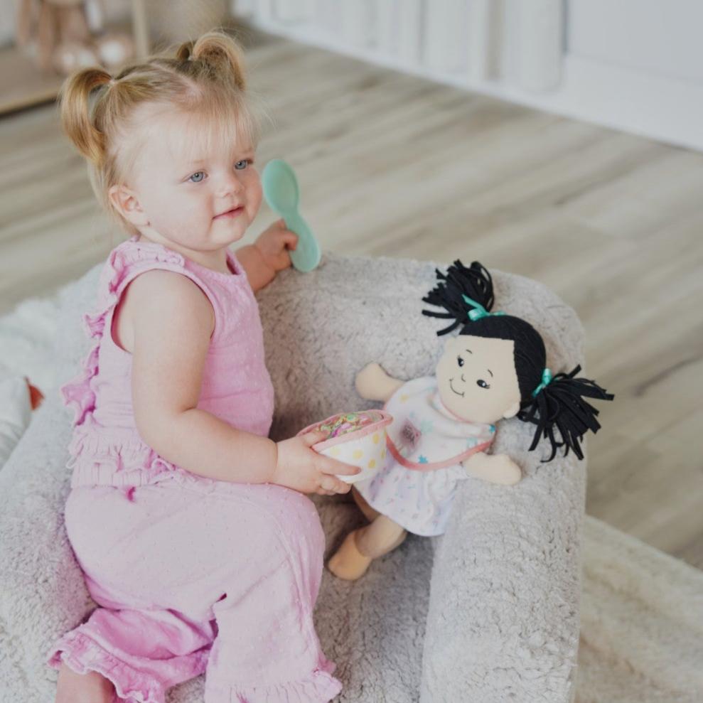 toddler age girl seated in a small gray chair holding a pretend food bowl and spoon with a soft doll nearby