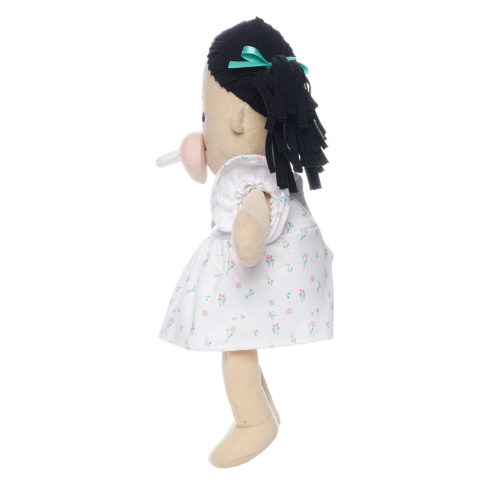 side profile of light beige soft doll with black pigtails in a dress seated with pink magnetic pacifier attached to the face