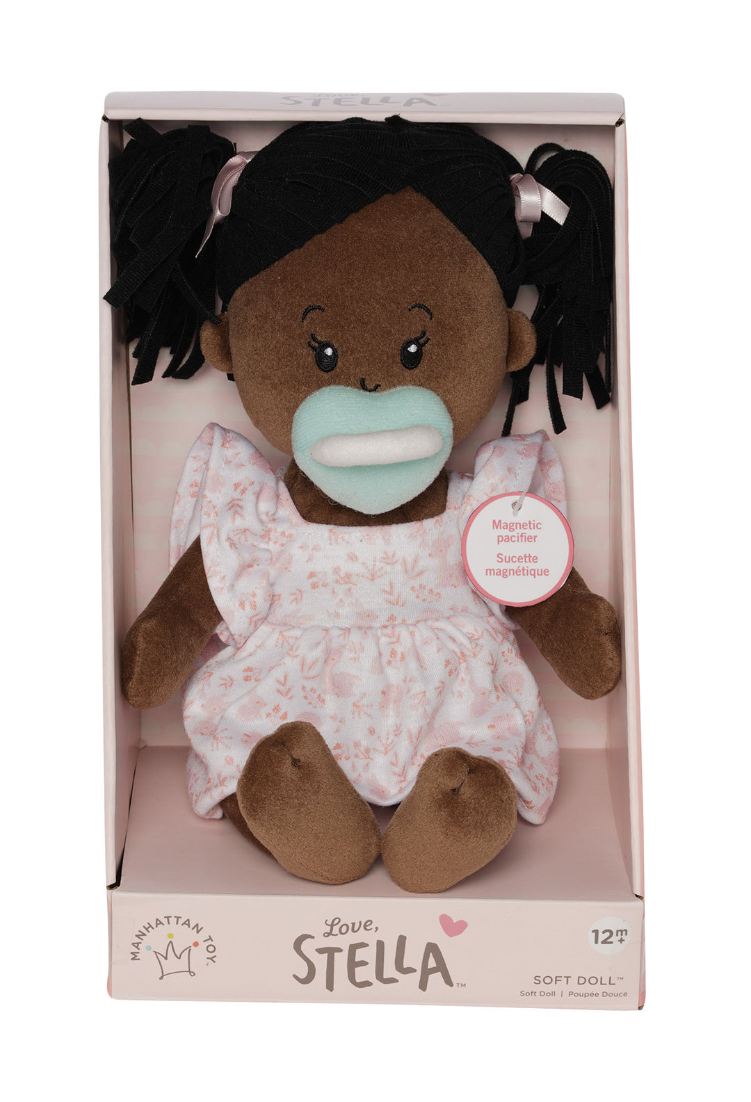 soft baby doll with brown skin and black hair wearing a dress with magnetic pacifier in open front box packaging