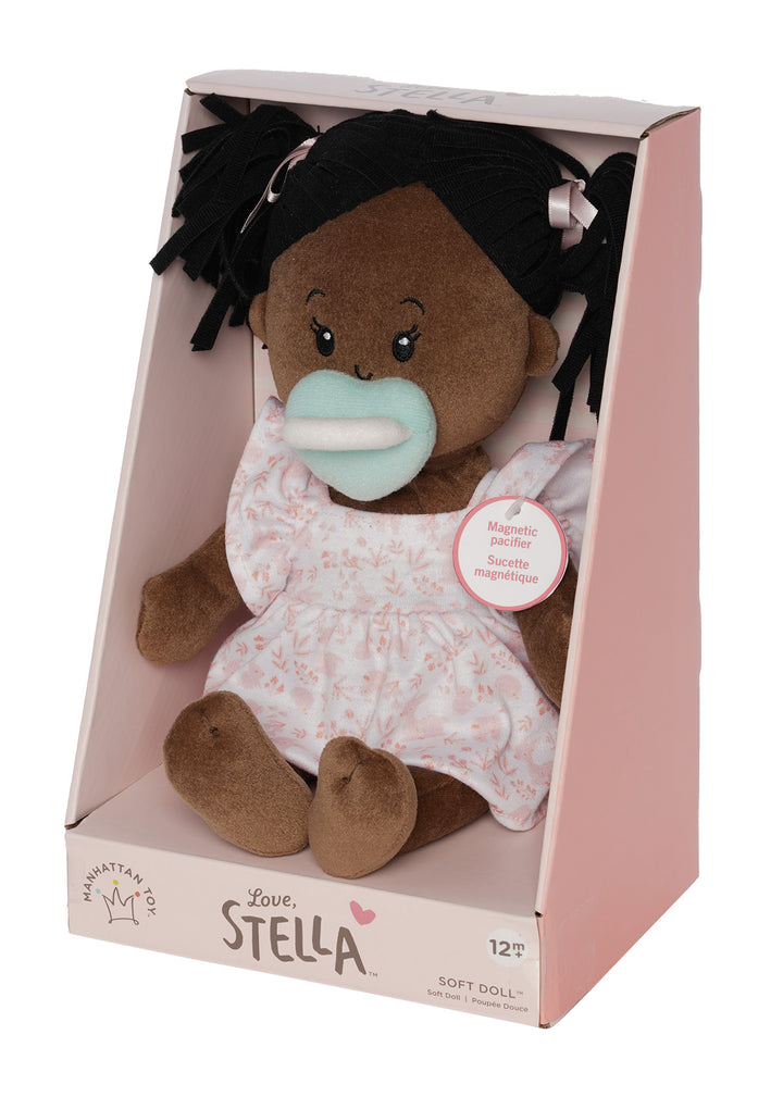 soft baby doll with brown skin and black hair wearing a dress with magnetic pacifier in open front box packaging