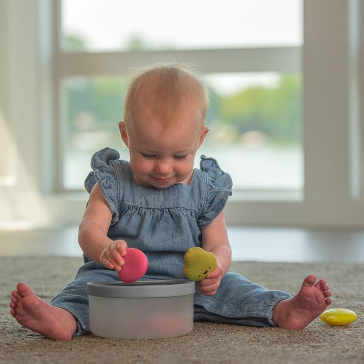 toddler sitting on the floor, smiling, playing with fruit fill n' spill toy