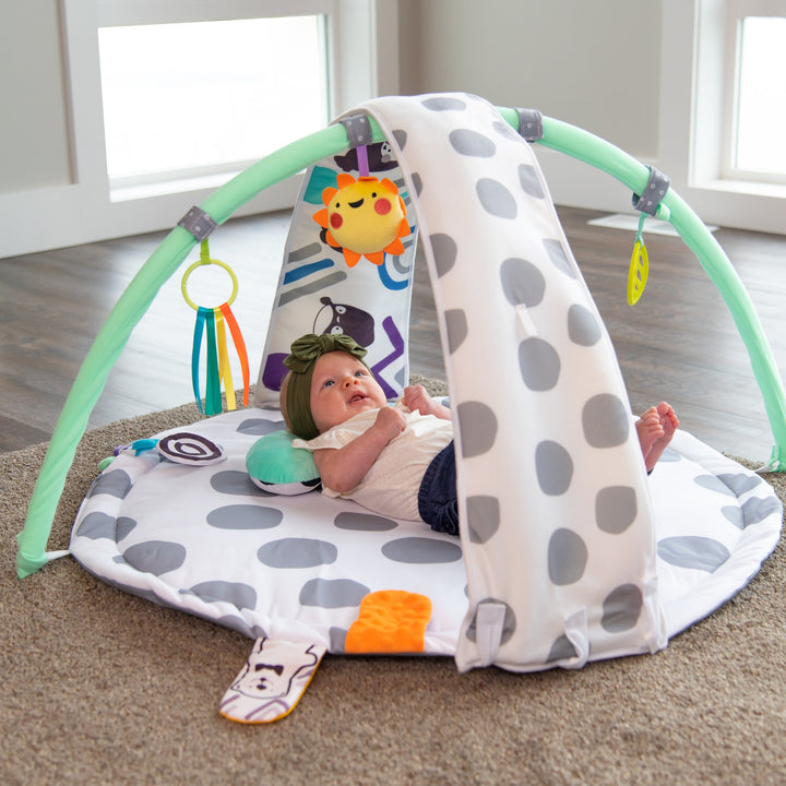 playmat with gray polka-dots, an arch with toys on it, a bolster, and floor mirror