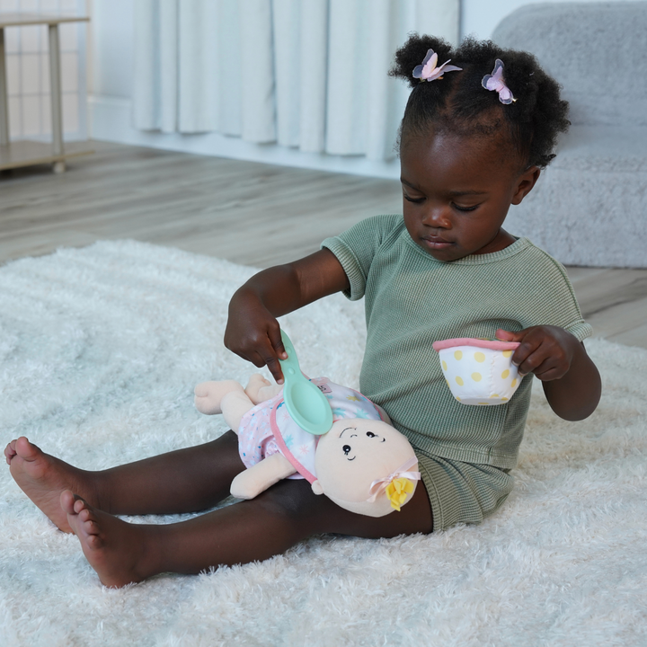 toddler age girl seated on the floor pretending to feed her soft baby doll with a pretend food bowl and plastic spoon